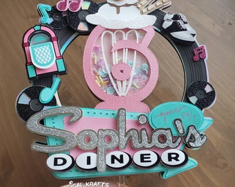 May include: A pink and teal diner-themed cake topper with a milkshake, musical notes, and a jukebox. The topper features the name "Sophia's Diner" in silver glitter and the words "open 24/7".