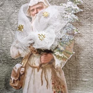 May include: A Christmas ornament featuring a vintage illustration of a figure in a white robe and a white head covering adorned with gold stars. The figure carries a small tree and a satchel. A clear ribbon is attached for hanging.