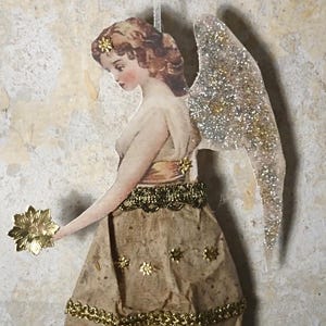 Puede incluir: Adorno de ángel de estilo vintage con una estrella dorada. El ángel tiene un vestido de papel con adornos dorados y alas brillantes. El pelo del ángel es castaño y sostiene una estrella dorada.