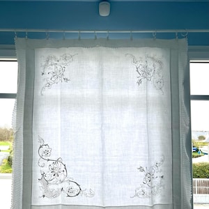 May include: White linen curtain with embroidered floral corner designs. The curtain has a fringed edge and is hanging in front of a window. The curtain is made of a sheer fabric, allowing light to pass through.