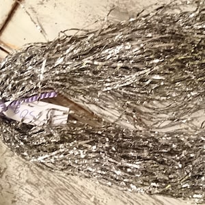 May include: A bundle of silver tinsel, tied with a purple and white cord. A white tag with handwritten text is attached. The tinsel is arranged in a circular shape, suggesting it may be a decorative item for the holidays.