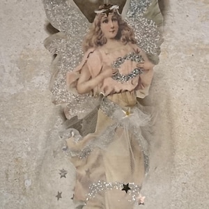 Puede incluir: Adorno de ángel de estilo vintage con alas cubiertas de purpurina y un vestido fluido. La figura del ángel está adornada con un halo y sostiene una corona. Los detalles de purpurina plateada y las estrellas añaden brillo.