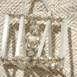 May include: A decorative hair accessory featuring a geometric design. The piece is composed of silver-colored beads and clear, rectangular plastic rods. A central cluster of beads includes a light-colored, round bead.