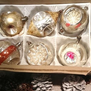 May include: A vintage box of six Christmas ornaments. The ornaments are various shapes and sizes, with silver and gold finishes. Some have red or pink accents. The box is cardboard and lined with white paper. Pine cones are scattered around the box.