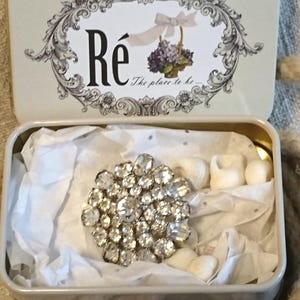 Pode incluir: Uma lata retangular aberta com uma tampa decorativa com a palavra "Ré" e um desenho floral. No interior, um grande broche de strass brilhante repousa sobre papel de seda. A lata é verde claro com uma borda dourada.