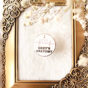 May include: A white enamel pin with a pink heart and the text "I ♥ GREY'S ANATOMY" inside a circle with a heartbeat line. The pin is displayed in a gold ornate frame.