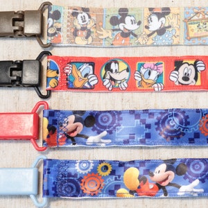Può includere: Quattro diversi fermagli in tessuto colorati con vari disegni di personaggi Disney, tra cui Topolino, Paperino e Pippo. I fermagli sono attaccati a uno sfondo bianco.