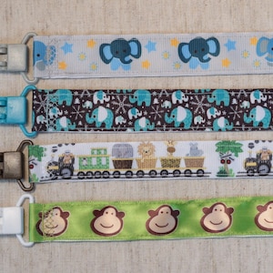 以下が含まれることがあります： 4種類のベビー用おしゃぶりクリップ。最初のクリップは、薄いグレーと白のリボンに青い象と黄色の星が描かれています。2番目のクリップは、茶色と青のリボンに青い象と白い雪の結晶が描かれています。3番目のクリップは、白いリボンに緑の電車と動物が描かれています。4番目のクリップは、緑のリボンに茶色の猿が描かれています。