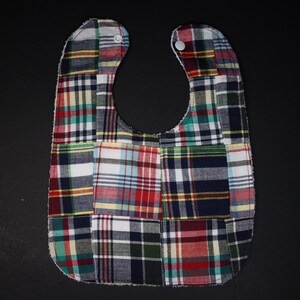 May include: A patchwork baby bib with a red, blue, and green plaid pattern. The bib has two white snaps on the neck.