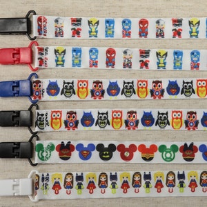 May include: Six colorful ribbon clips with different cartoon characters. The clips are black, white, red, and blue. The characters include superheroes, owls, Mickey Mouse, and girls.