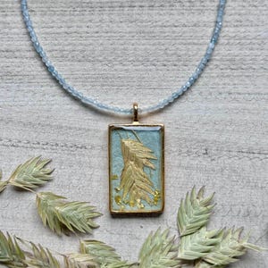 May include: A necklace with a rectangular pendant showcasing a gold-coloured leaf design on a light blue backdrop. The pendant hangs from a strand of small, light blue beads. Dried green and tan leaves are arranged around the necklace.