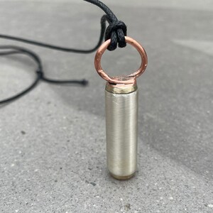 May include: A silver and copper pendant necklace with a cylindrical charm. The charm has a smooth, brushed finish and is attached to a simple copper ring.