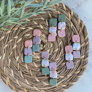 Könnte beinhalten: Ein Set mit sechs Paaren Hängeohrringen mit einem mehrschichtigen Design. Jeder Ohrring besteht aus drei quadratischen Tonperlen in den Farben Rosa, Grün und Lila. Die Perlen sind mit goldfarbenen Metallakzenten verziert. Die Ohrringe werden auf einer geflochtenen Weidenoberfläche präsentiert.