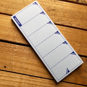 May include: A white weekly planner notepad with blue corner tabs labeled with the days of the week: Mon, Tue, Wed, Thu, Fri, Sat, Sun. The notepad is on a wooden surface.