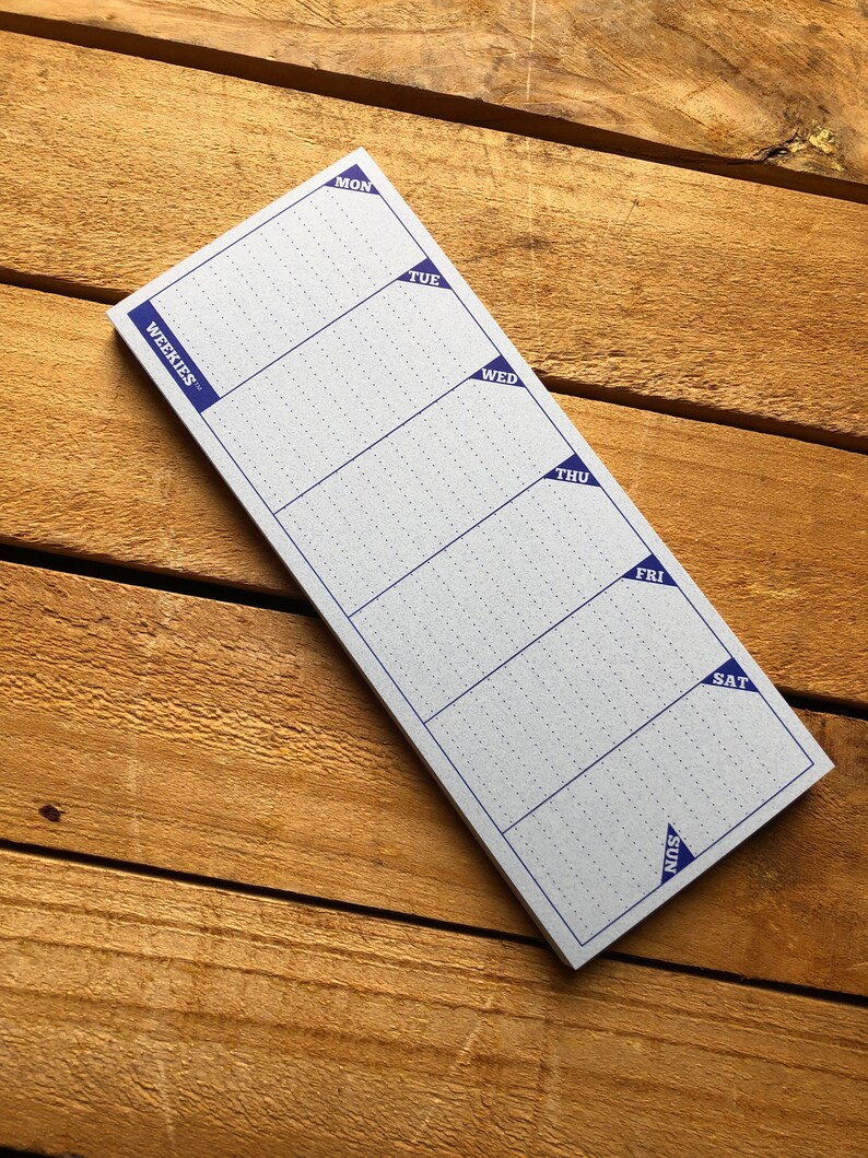 May include: A white weekly planner notepad with blue corner tabs labeled with the days of the week: Mon, Tue, Wed, Thu, Fri, Sat, Sun. The notepad is on a wooden surface.