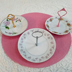 May include: Three white ceramic cake stands with floral designs. Each stand has a metal handle and is perfect for serving desserts or snacks.