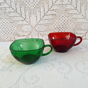 May include: Two square-shaped glass teacups, one green and one red, with handles. The cups are sitting on a white crocheted tablecloth.