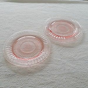 May include: Two pink glass dessert plates with a scalloped edge and a sunburst pattern in the center.