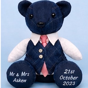 May include: A navy blue teddy bear dressed in a white shirt, a plaid waistcoat, and a pink tie. The bear's paws are embroidered with "Mr & Mrs Askew" and "21st October 2023". The bear has black eyes and a black nose.