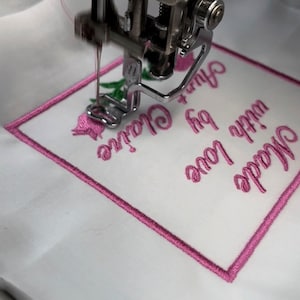 May include: Close-up of a pink embroidery machine stitching a pink square with the text "Made with love + a little bit of magic" on a white fabric.