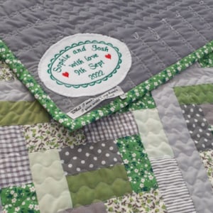 May include: A grey and green patchwork quilt with a white oval label that reads "Sophie and Josh with love 9th Sept 2022". The label has two red hearts.