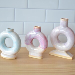 May include: Three ceramic vases with a ring-shaped opening in the middle. The vases are glazed in a light blue, pink, and purple colour.