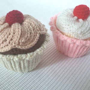 Könnte beinhalten: Zwei handgestrickte Cupcakes. Einer hat eine hellbraune Oberseite und eine weiße Basis, der andere hat eine weiße Oberseite und eine rosa Basis. Beide haben eine rote Kirsche oben drauf. Die Cupcakes stehen auf einer weißen Oberfläche.