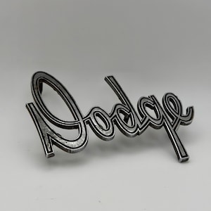 May include: A silver-toned metal Dodge emblem featuring the brand name in a stylized, cursive script font. The letters have a raised, outlined design with a dark, recessed interior, creating a dimensional appearance for this automotive badge.