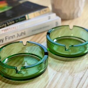 May include: Two round, translucent green glass ashtrays, each featuring three evenly spaced notches along the rim for resting items. The ashtrays have a smooth, polished finish and a thick, circular base, showcasing a vibrant emerald green hue.