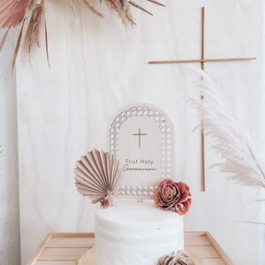May include: A white cake with a wooden topper that says "First Holy Communion" and a cross. The cake is decorated with dried flowers and a fan-shaped paper decoration.