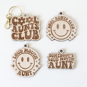 May include: Four wooden keychains with different designs. One keychain features the text "Cool Aunts Club" in a retro font with a flower design. Another keychain features a smiley face with the text "Cool Aunts Club" around it. The third keychain features a smiley face with the text "Loud Mouth Aunt" around it. The fourth keychain features the text "Somebody's Loud Mouth Aunt" in a retro font.