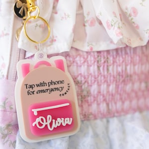 May include: A pink backpack-shaped keychain with the name "Olivia" in white script. The keychain has the text "Tap with phone for emergency contact". The background features a floral pattern and a gold-colored clasp.