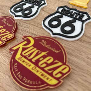 May include: Two white and black Route 66 shield-shaped signs with raised lettering, two yellow translucent trophy-shaped cutouts, and two circular red and yellow Rust-eze Bumper Ointment Rear End Formula signs with raised text and graphics.
