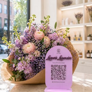 May include: A bouquet of lavender and pink flowers wrapped in brown paper, with a light purple sign that reads "Lamerè Lavender" and includes a QR code. The sign is on a marble countertop.