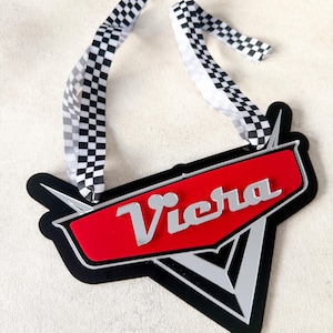 May include: A black, red, and silver Viera logo with a checkered ribbon. The logo has a red background with the word "Viera" in silver. The ribbon is black and white checkered.