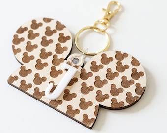 May include: A wooden keychain shaped like Mickey Mouse ears with a brown Mickey Mouse pattern. The keychain has a gold metal ring and a gold metal clasp. A white plastic letter "J" is attached to the keychain.