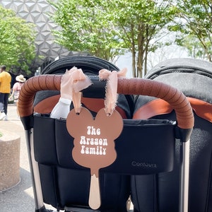 May include: A brown Mickey Mouse-shaped popsicle with the text "The Arason Family" in white. The popsicle is attached to a black stroller with brown leather handles.