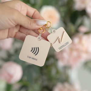 May include: Two square-shaped tags with gold rings. One tag has a contactless payment symbol and the text "TAP HERE". The other tag has a handwritten "N" and the text "YANESSA VIERA PHOTOGRAPHY".