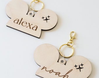 May include: Two wooden keychains in a film camera shape, with gold-colored hardware. One keychain has the name "alexa" engraved, and the other has "noah". Each keychain has a starburst design.