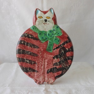 May include: A ceramic cat figurine with a red and black striped body, a white face, and a green bow with blue polka dots. The cat has yellow eyes and a black nose. The figurine is a decorative item.