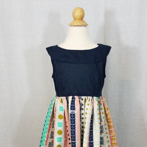 Puede incluir: Vestido sin mangas azul marino con una falda estampada colorida. La falda presenta rayas verticales en rosa, blanco y turquesa, adornadas con formas geométricas y animales. El vestido se exhibe en un maniquí.