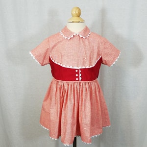 Puede incluir: Un vestido de estilo vintage con un patrón rojo claro y blanco, con un corpiño rojo y una falda acampanada. El vestido tiene un cuello y bordes de manga festoneados, con botones y ribetes blancos. El vestido se exhibe en un maniquí de madera.