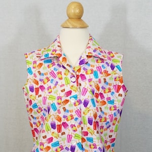 May include: Sleeveless button-down shirt with a white background covered in colorful popsicle illustrations. The shirt has a collared neckline and a pocket. The popsicles are in shades of red, orange, blue, green, and purple.