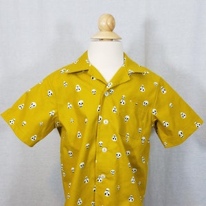 Puede incluir: Camisa de manga corta de color amarillo mostaza con un estampado de calaveras blancas. La camisa tiene un cuello clásico, un bolsillo en el pecho y botones blancos. La camisa se muestra en un maniquí.
