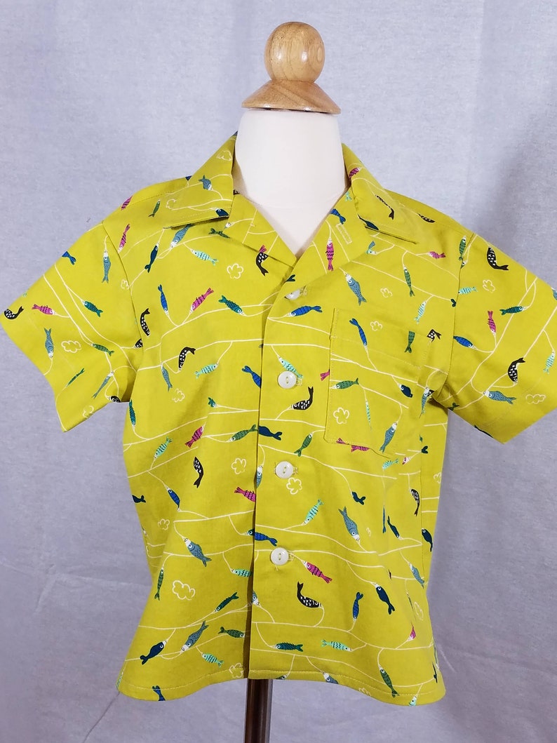 May include: A short-sleeved, button-down shirt in a bright yellow-green color. The shirt features a repeating pattern of colorful fish and white wavy lines. It has a chest pocket and white buttons.