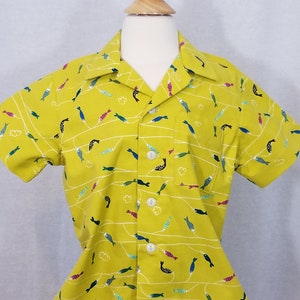 May include: A short-sleeved, button-down shirt in a bright yellow color, adorned with a repeating pattern of colorful fish and white wavy lines. The shirt has a single chest pocket and white buttons. The shirt is displayed on a mannequin.