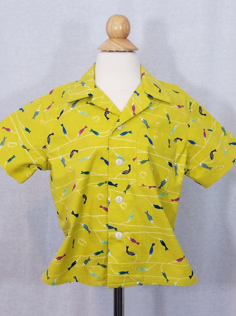 May include: A short-sleeved, button-down shirt in a bright yellow color, adorned with a repeating pattern of colorful fish and white wavy lines. The shirt has a single chest pocket and white buttons. The shirt is displayed on a mannequin.