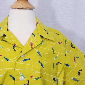 May include: A short-sleeved button-up shirt in a bright yellow-green color. The shirt features a repeating pattern of colorful fish and white lines. The shirt has a single pocket on the chest and white buttons.
