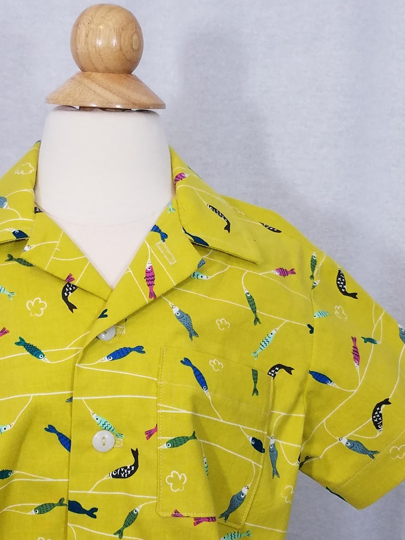 May include: A short-sleeved button-up shirt in a bright yellow-green color. The shirt features a repeating pattern of colorful fish and white lines. The shirt has a single pocket on the chest and white buttons.