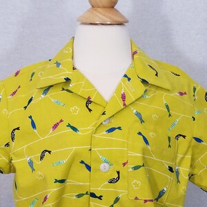 May include: A short-sleeved button-down shirt in a bright yellow-green color, adorned with a repeating pattern of colorful fish and white wavy lines. The shirt has a single chest pocket and white buttons.